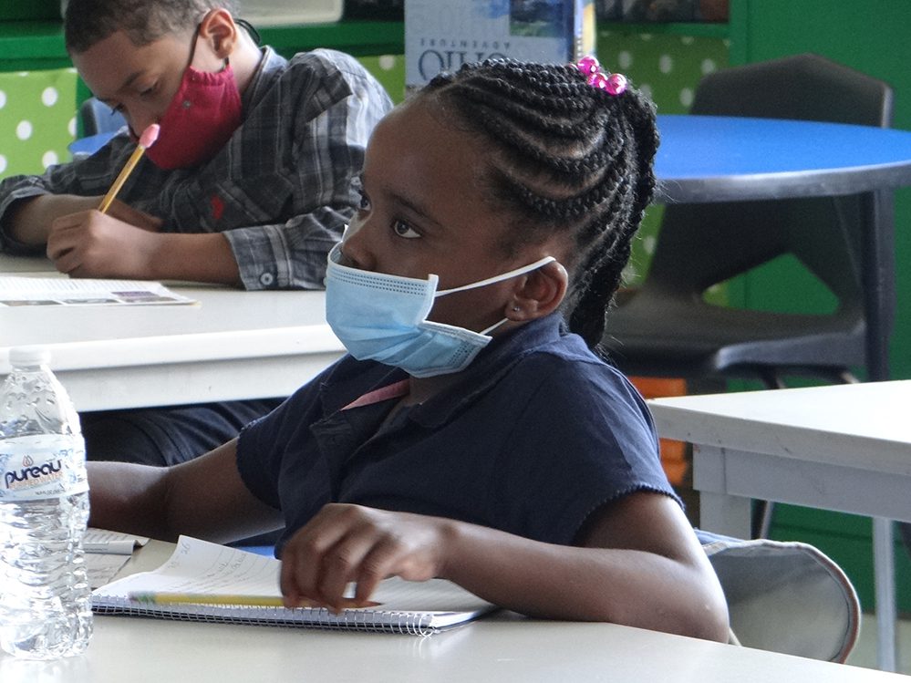 Two masked children at desks, one writing, one looking forward.