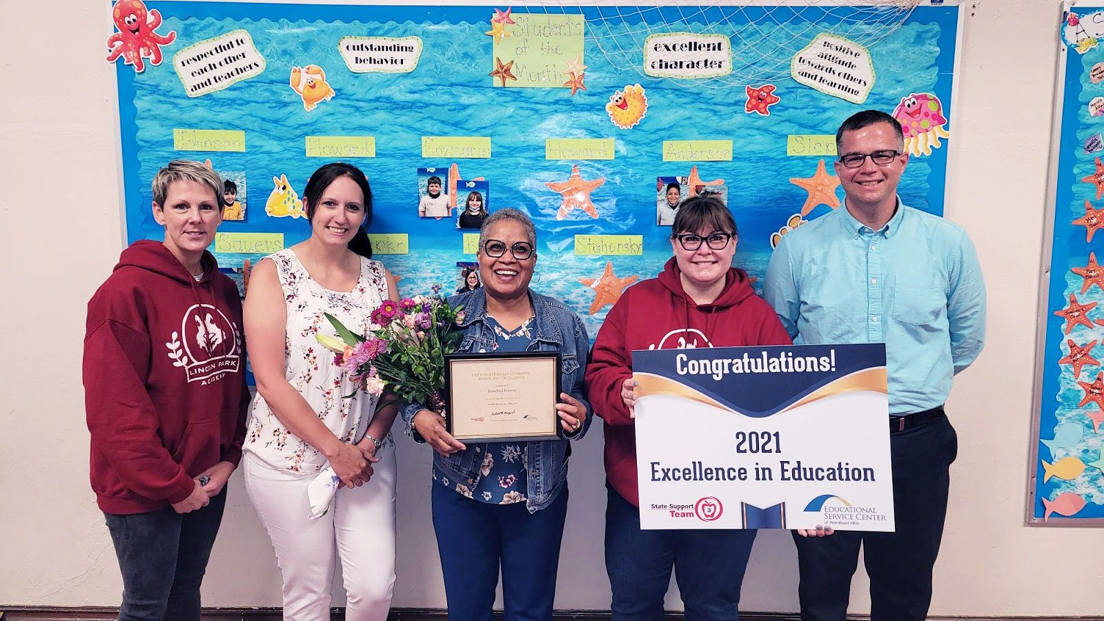 Five adults holding awards and flowers in front of a decorated bulletin board.