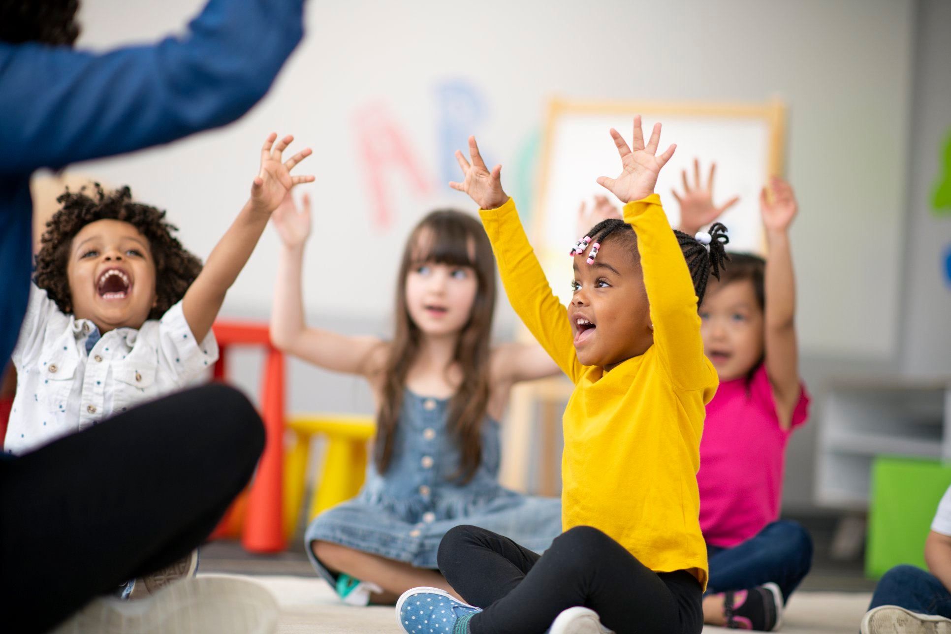 Young children sitting on floor with arms raised, participating in an activity.