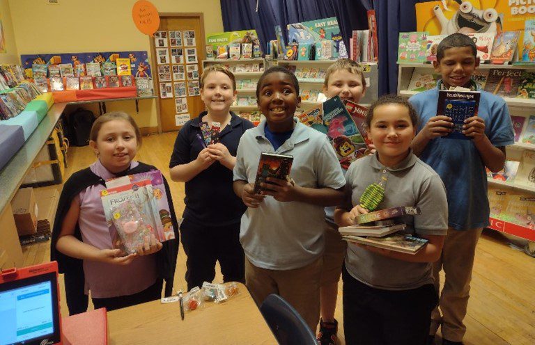 Six children smiling and holding books and items at a book fair.