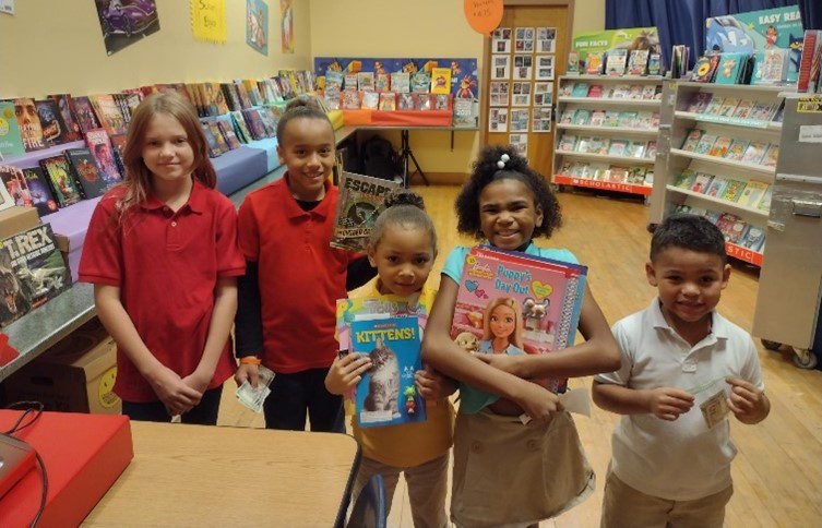 Five children smiling, holding books and money at a book fair.