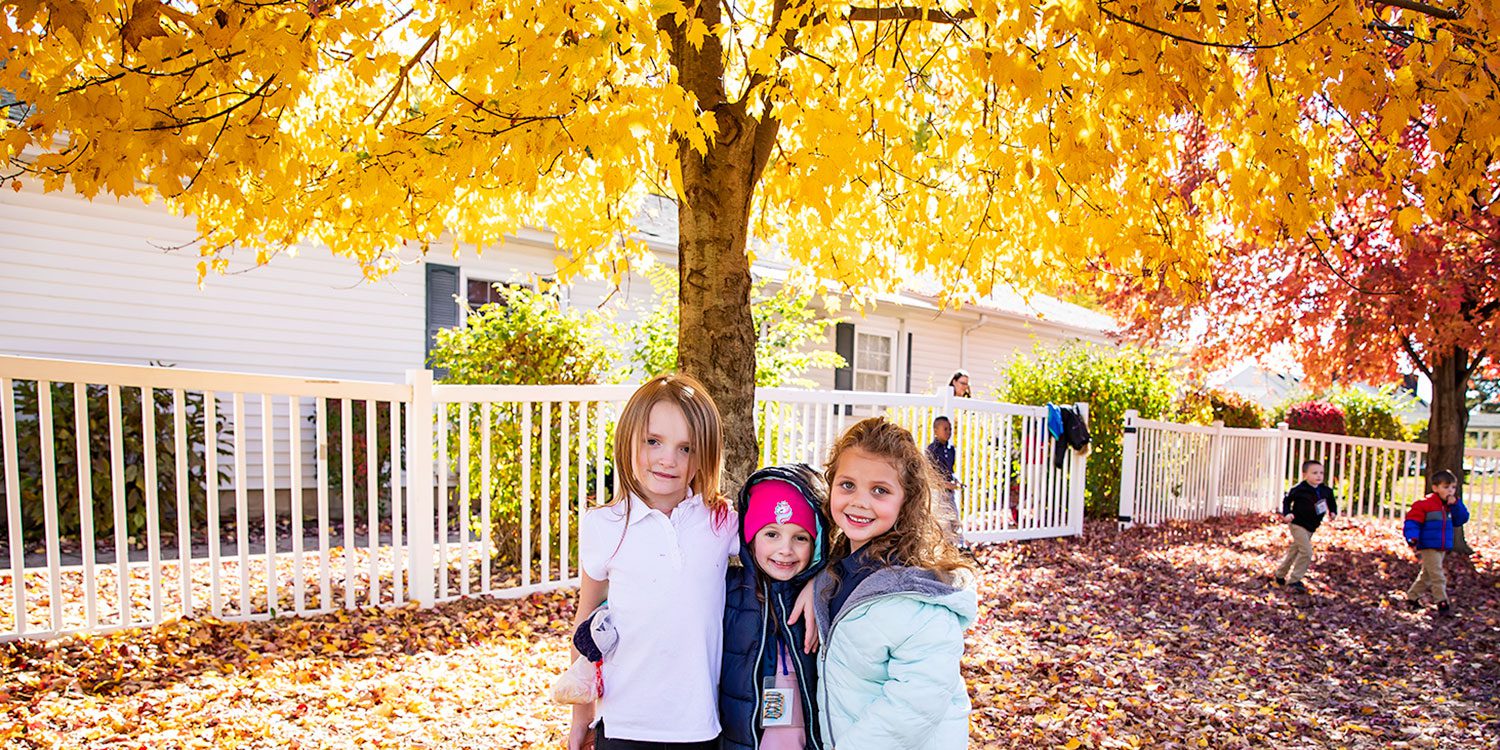 Three young children smiling under a tree with yellow autumn leaves.