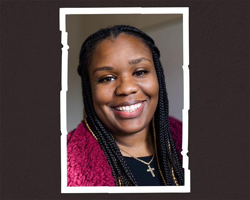 Smiling adult with braided hair and cross necklace in a photo frame.