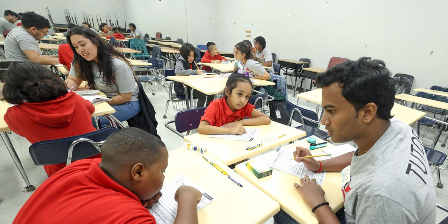 Adults and children work at desks in a brightly lit classroom.