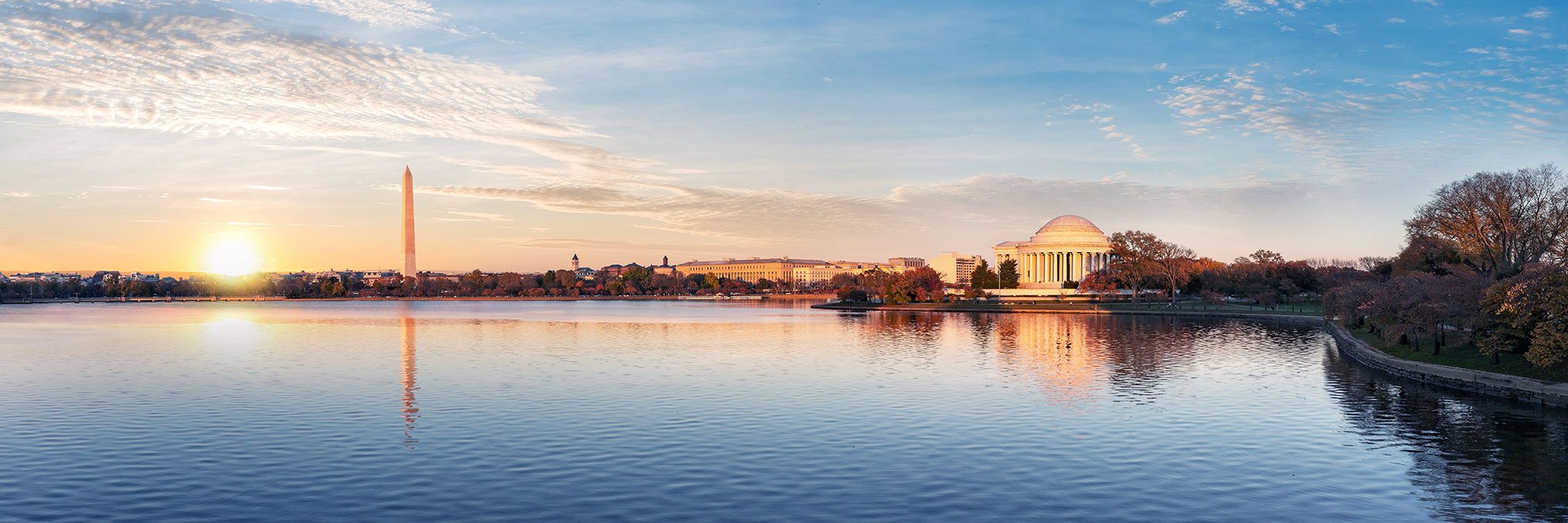 Sunrise over city skyline with obelisk and domed building reflected in water.