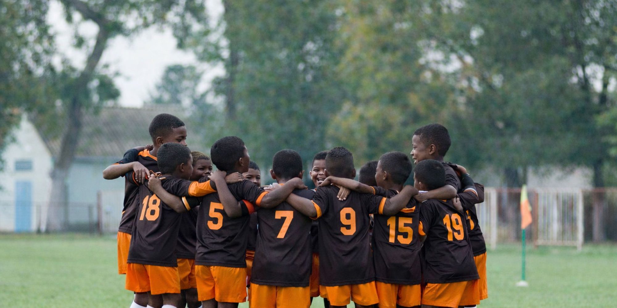 Children in soccer uniforms huddle on a field, arms around each other.