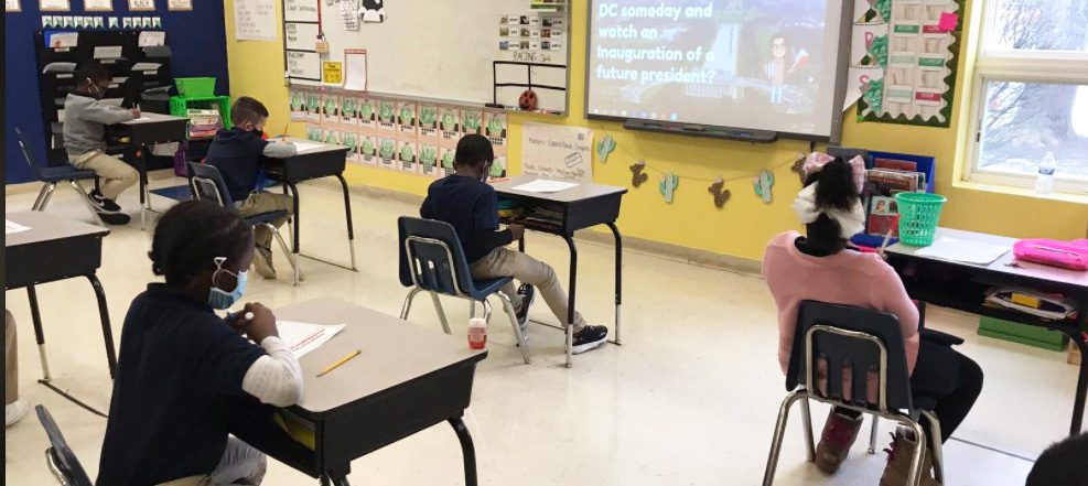 Children seated at desks in a classroom, looking towards a screen.