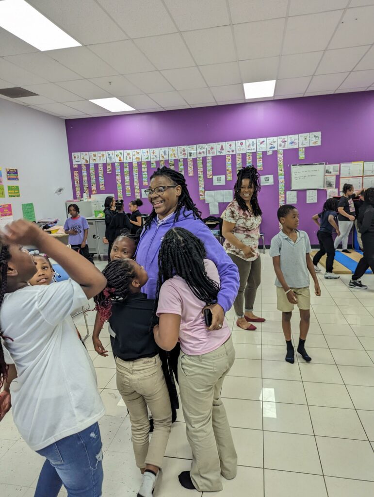 Smiling adult with dreadlocks surrounded by children in a room.
