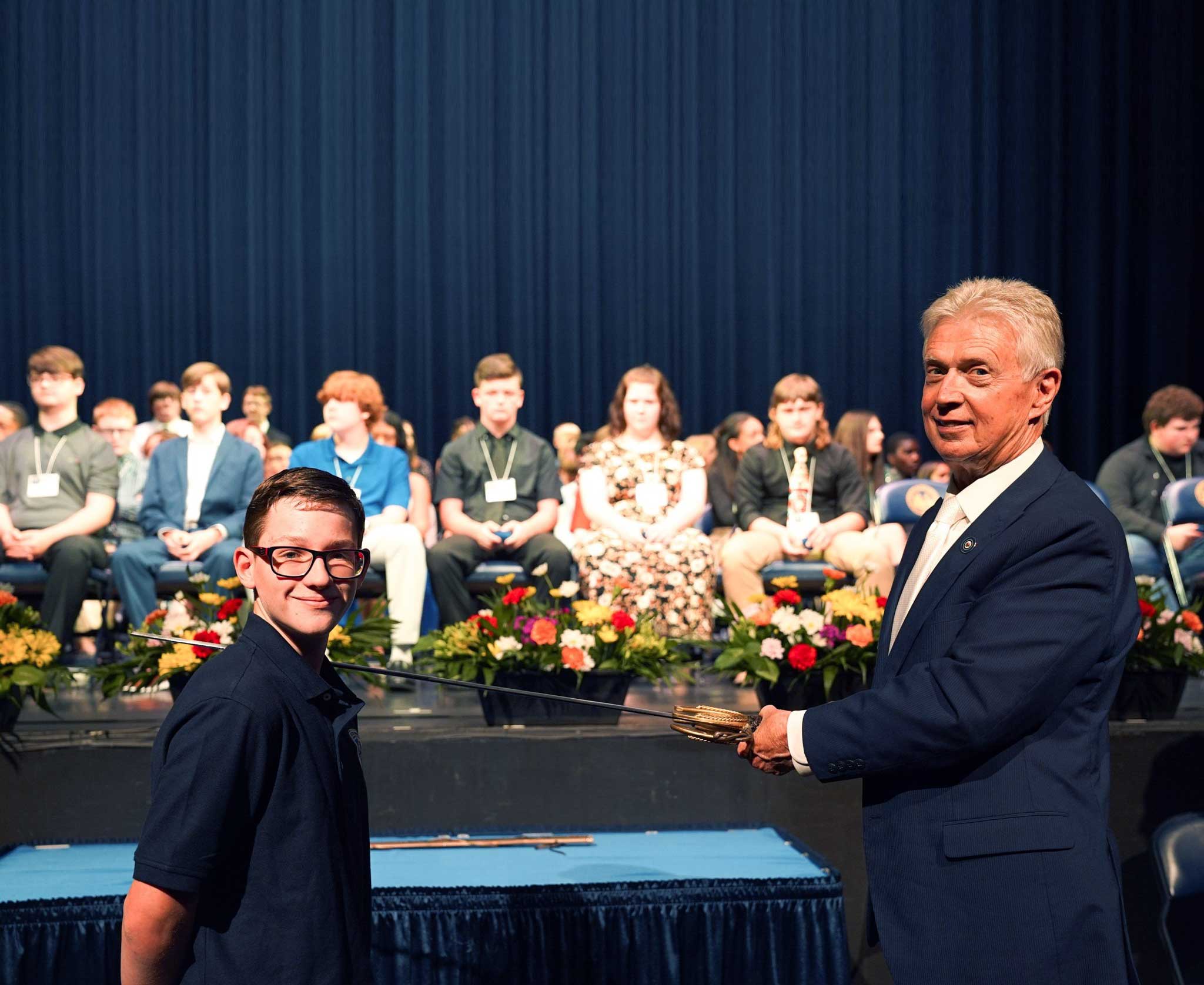 Adult presents ceremonial sword to child on stage.