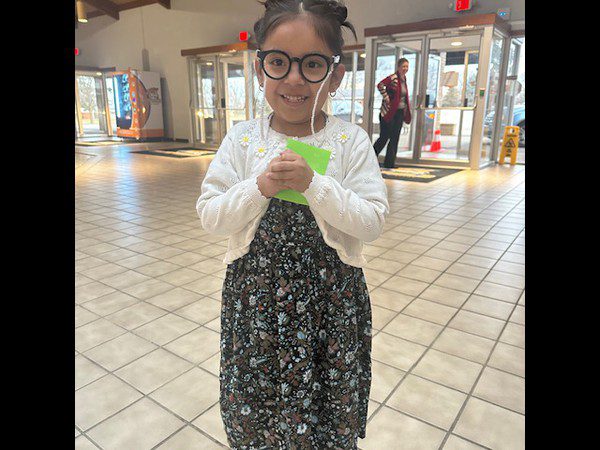 Smiling child in glasses holds green paper in a hallway.