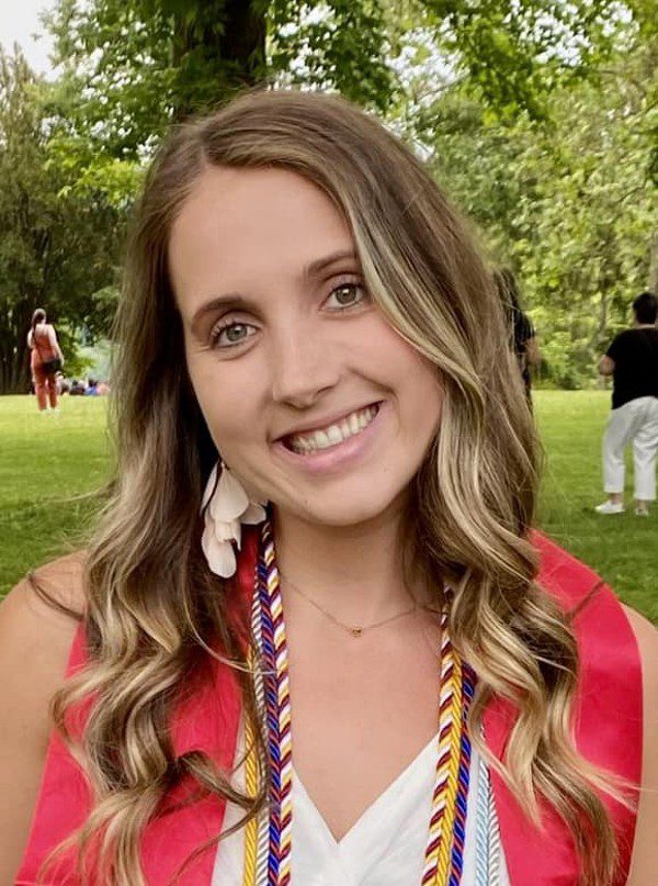 Smiling adult in graduation stole and cords outdoors.