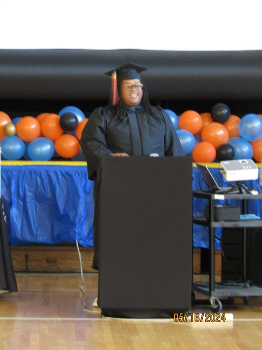 Adult in graduation cap and gown smiling behind a podium.