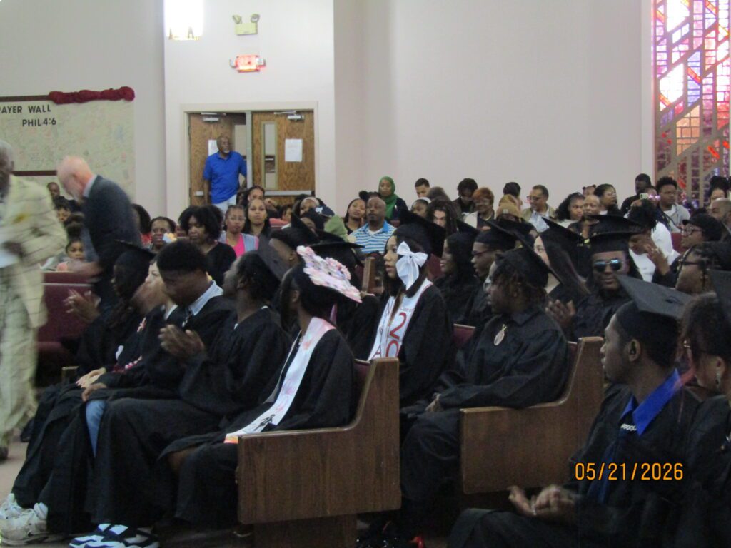 Audience, many in graduation attire, seated and clapping.