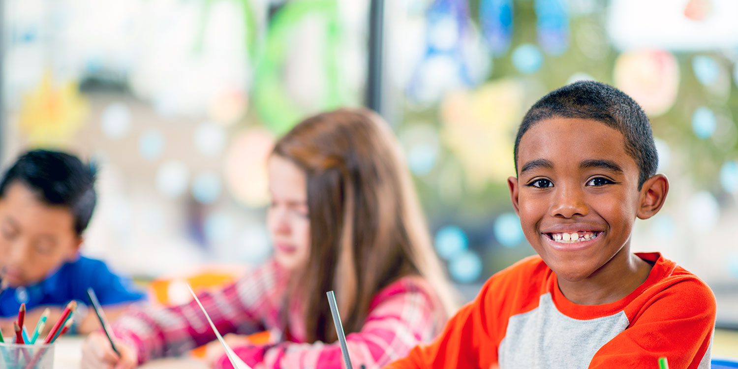 Smiling child in classroom with other children drawing.