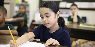 Child writing with a pencil at a desk in a classroom.
