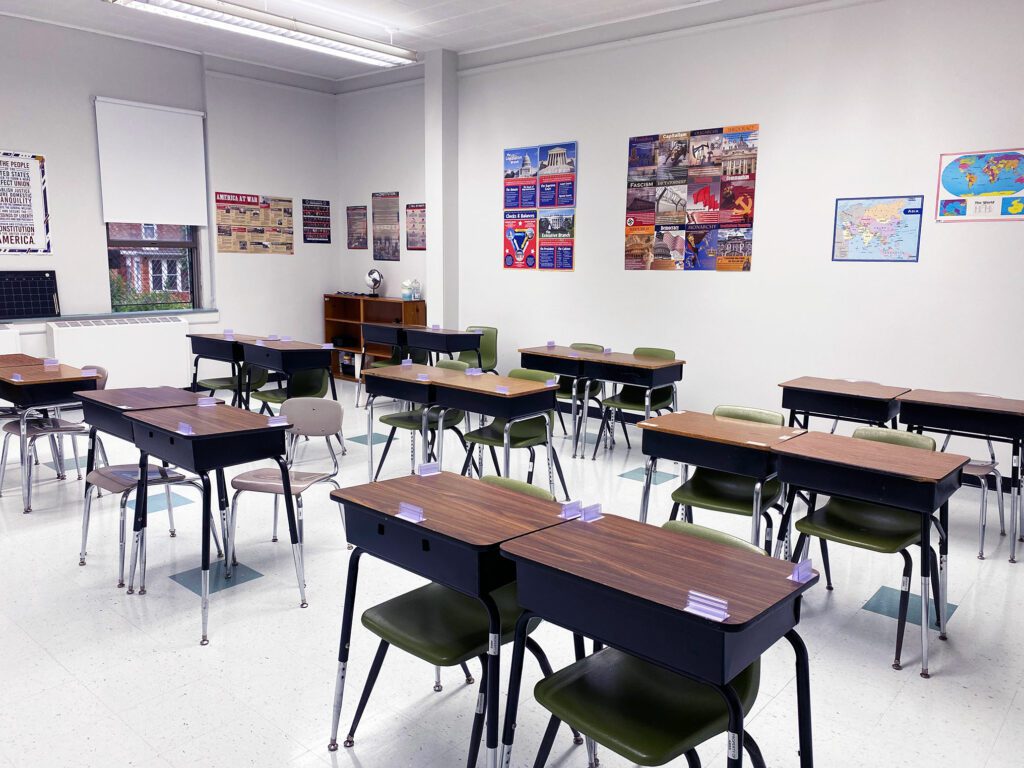 Empty classroom with student desks, chairs, and educational posters on white walls.