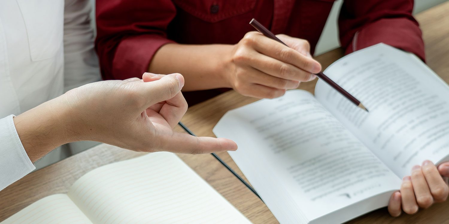Two people studying an open book with a pencil.