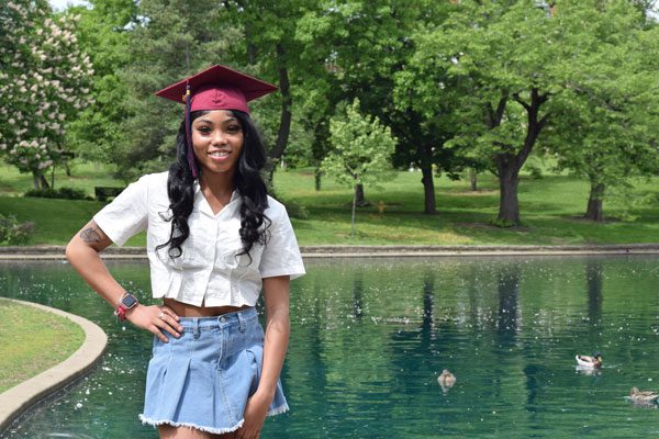 Adult in graduation cap smiling by a pond with ducks.
