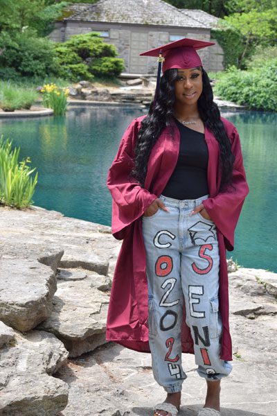 Adult in maroon graduation cap and gown by a pond.
