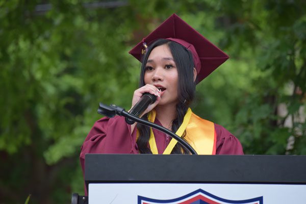 Adult in graduation attire speaking into a microphone at a podium.