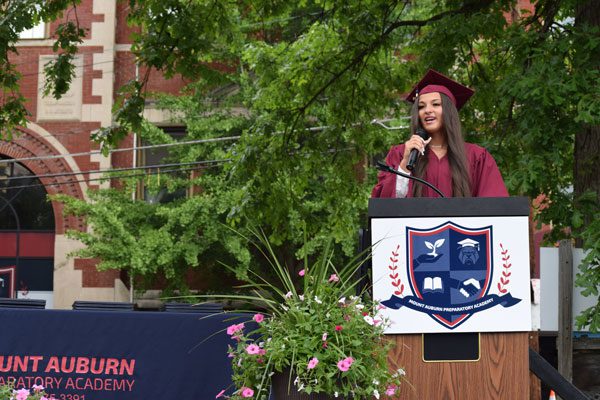 Adult in graduation cap and gown speaking at a podium.