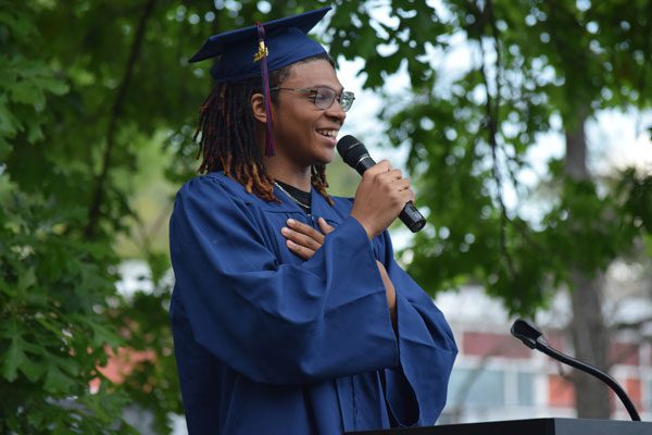 Adult in graduation cap and gown speaking into a microphone.