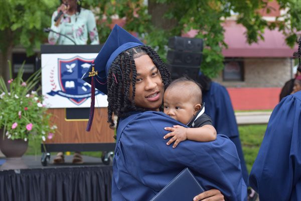 Adult in graduation attire holding a baby near a podium.