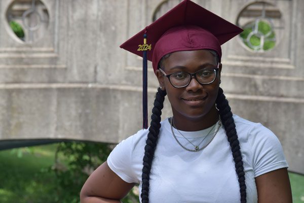 Young adult in maroon graduation cap and glasses smiles.