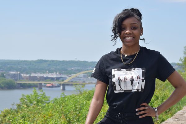 Adult smiling at camera with city, river, and bridge in background.