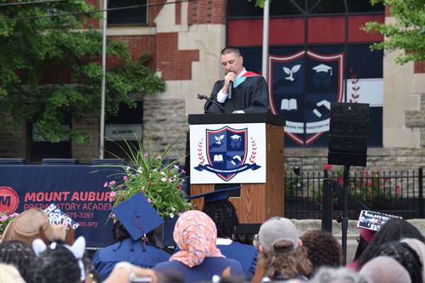 Adult in graduation attire speaking at a podium to an audience.
