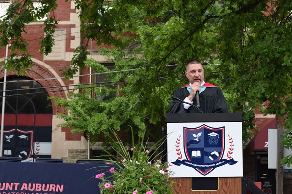 Adult in graduation attire speaking at podium with school crest.
