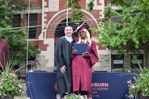 Two adults in graduation attire smiling and holding a diploma.