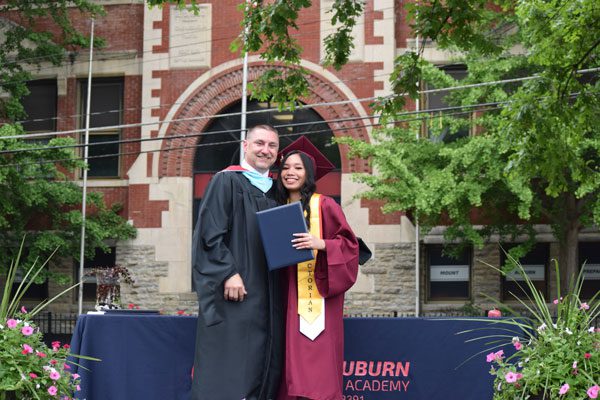 Two adults in graduation attire smiling, one holding a diploma.