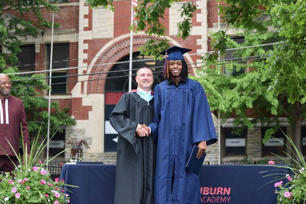 Two adults in graduation gowns shaking hands.