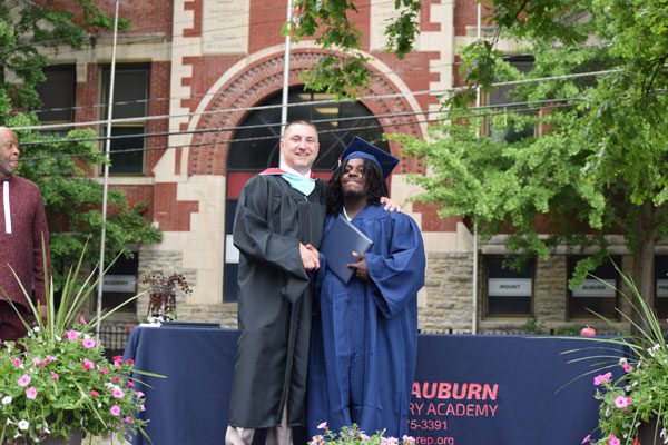 Two adults in graduation attire shake hands.