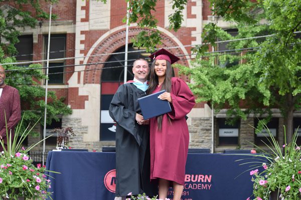 Two adults in graduation attire shake hands, one holding a diploma.