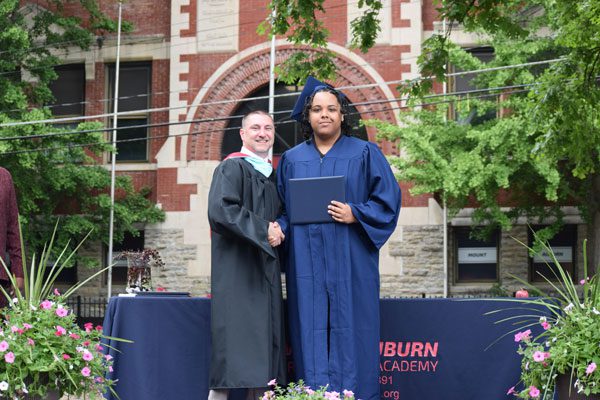 Two adults in graduation attire shake hands.