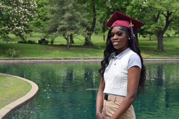 Smiling graduate in maroon cap and gown by a pond.