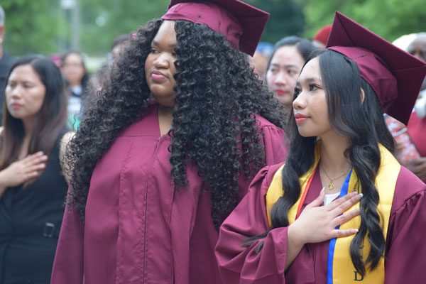 Two graduates in maroon caps and gowns with hands over hearts.