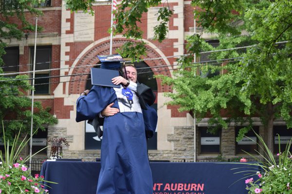 Two adults in graduation attire embrace on a stage.
