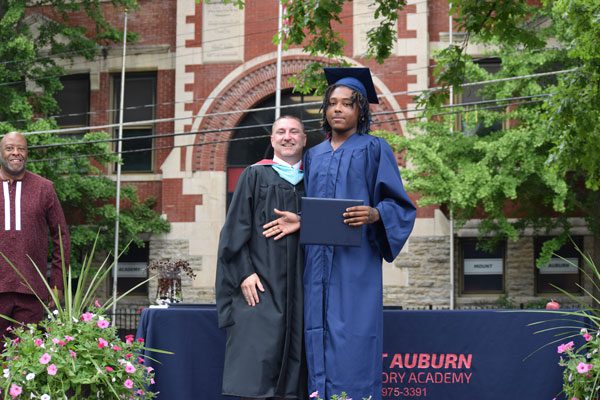 Two adults in graduation attire pose with a diploma.