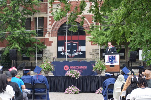 Speaker at podium addresses graduates and audience outdoors.
