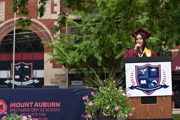Young adult in graduation attire speaking at a podium.