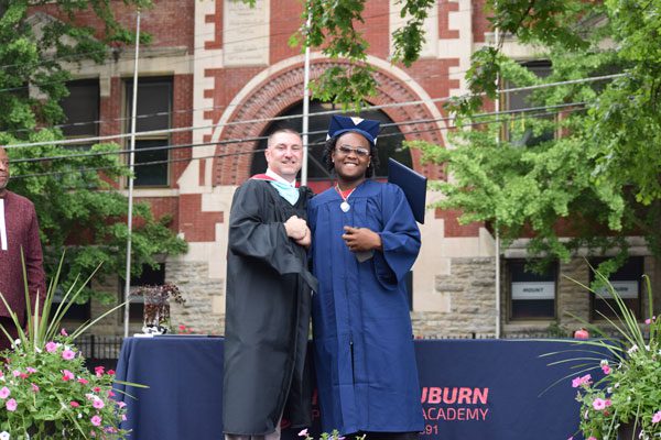 Two adults in graduation gowns pose outdoors.