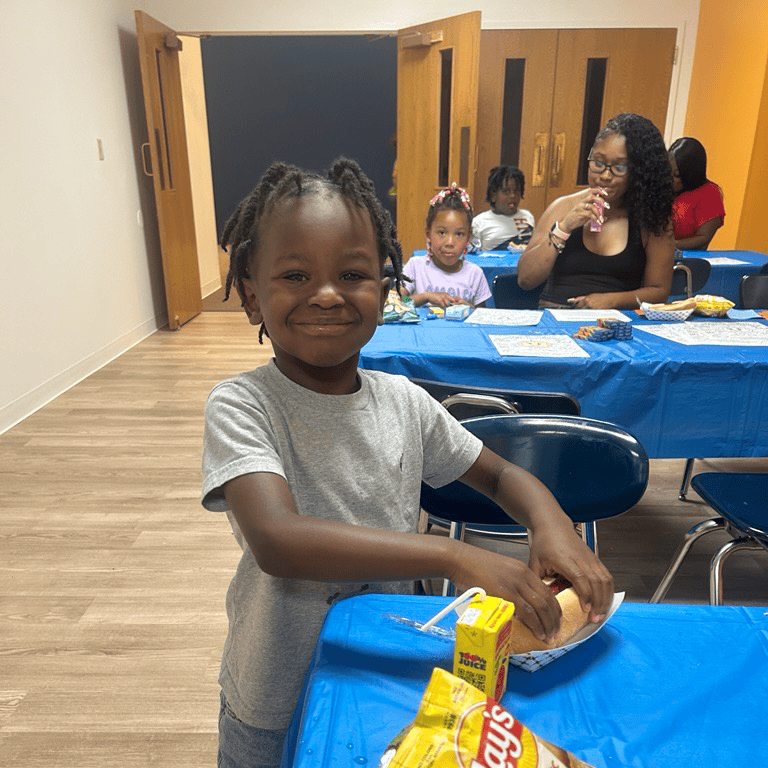 Child smiling, holding a hot dog bun at a table with snacks.