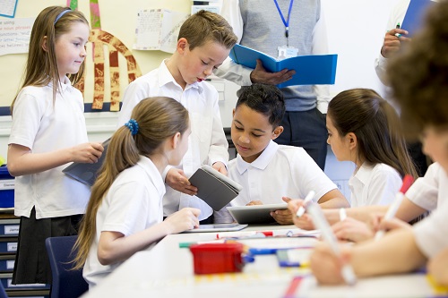Children in white shirts using tablets and collaborating in a classroom.