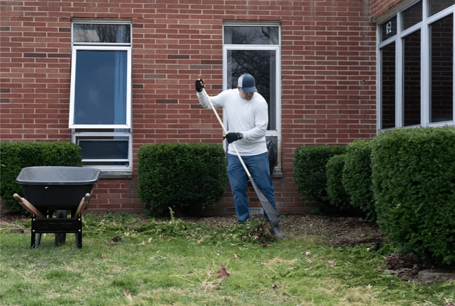 Adult raking leaves and debris next to a brick building.