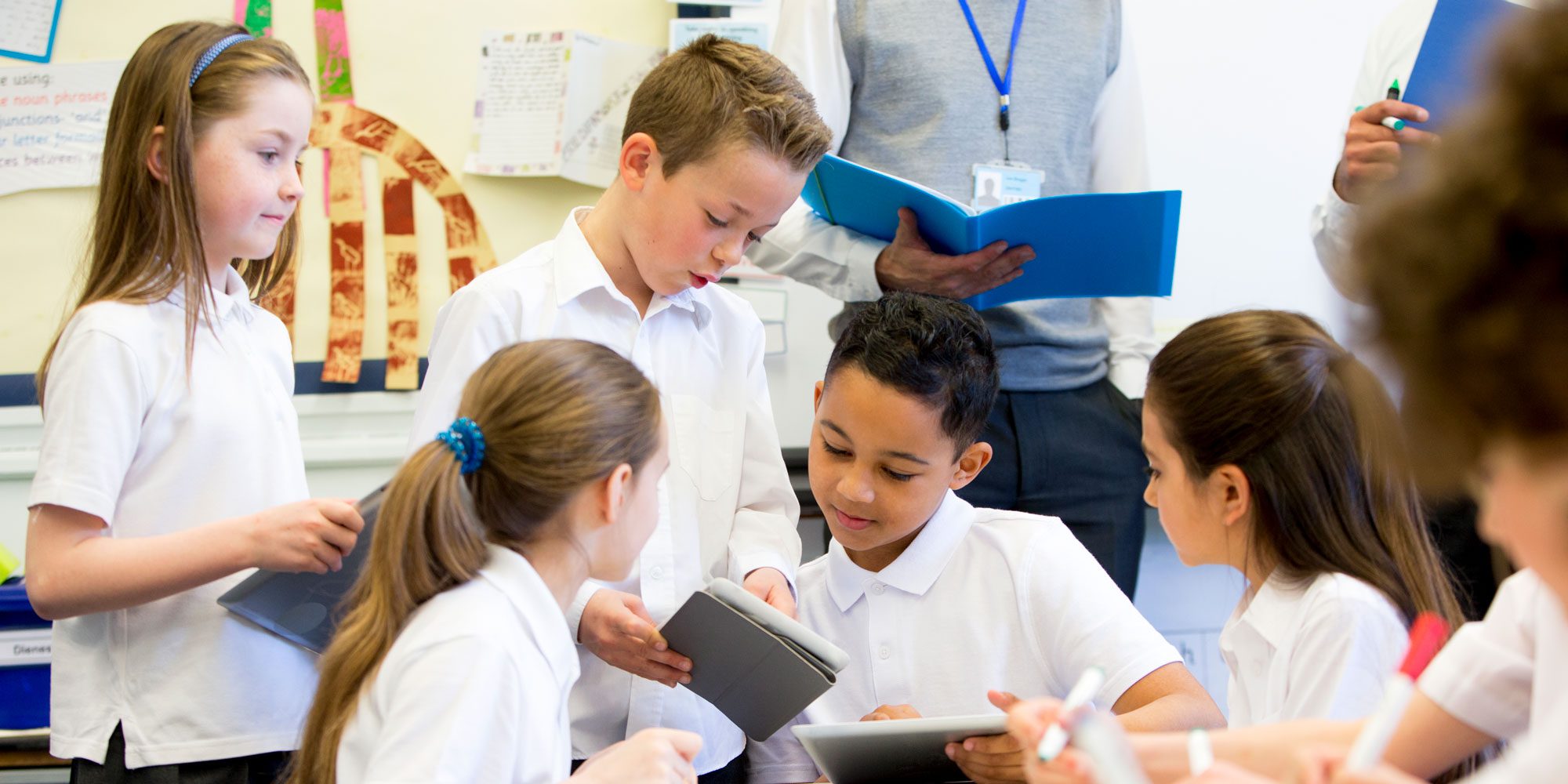 Children in white shirts using tablets in a classroom.