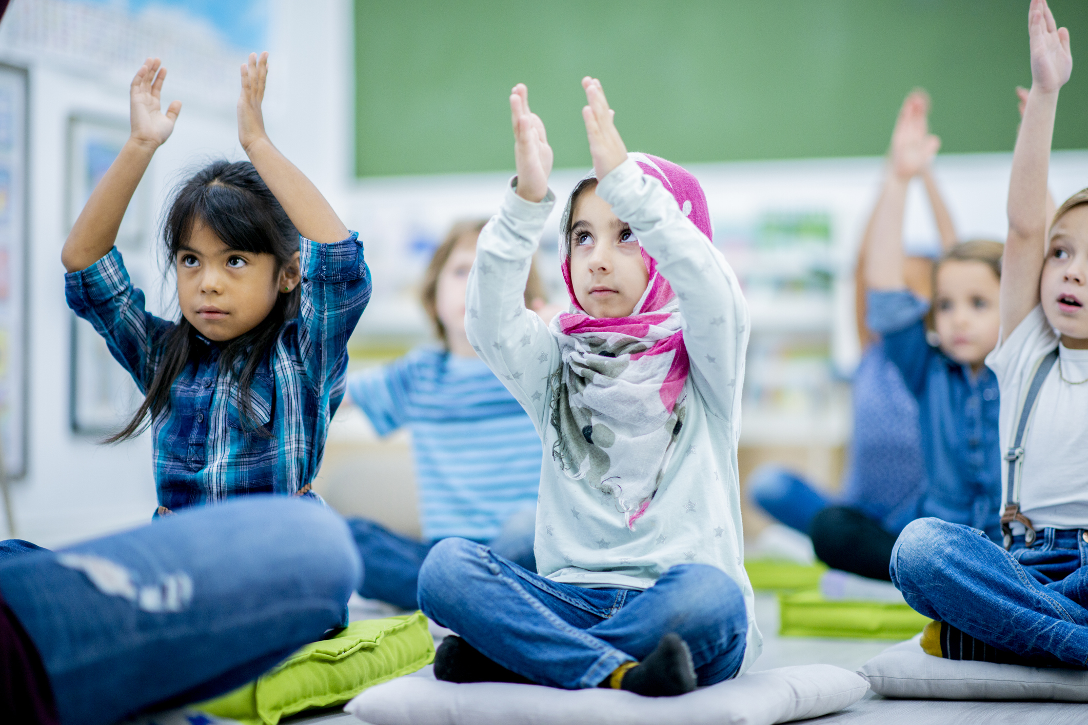Children sitting on cushions with hands raised in a classroom.