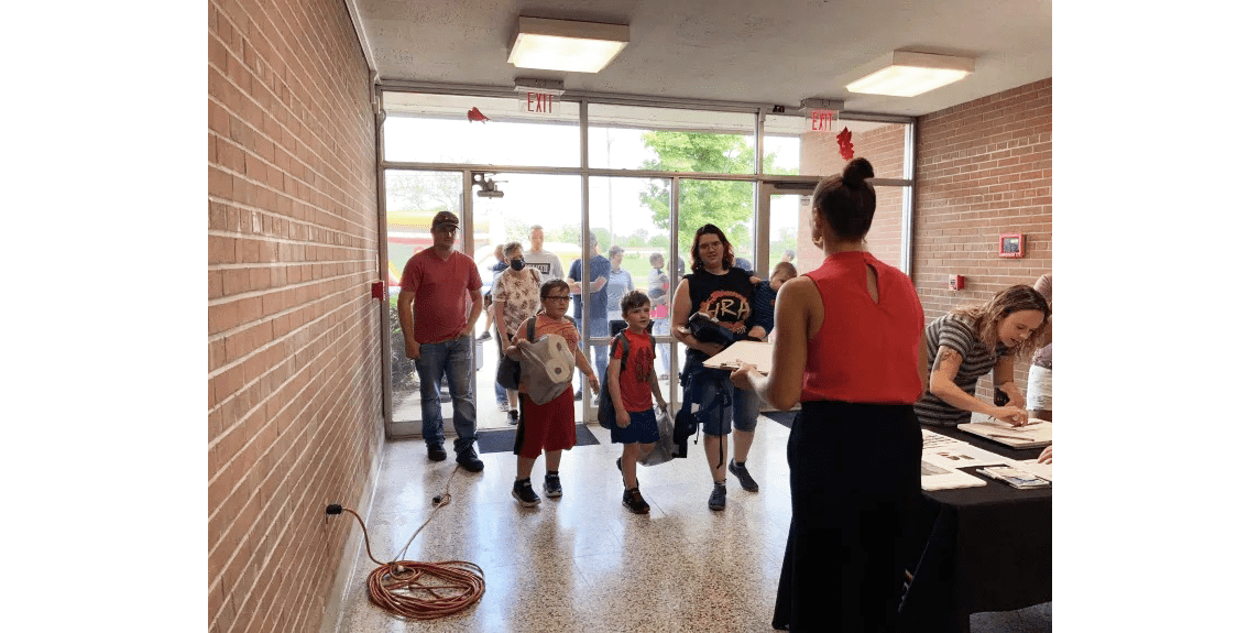 Adults and children entering a building, greeted by adults at a table.